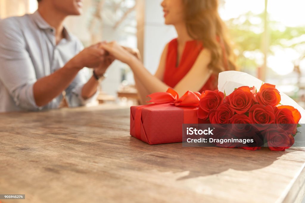 Celebrating Valentines Day with Soulmate Cheerful young couple sitting at wooden table of outdoor cafe and holding hands while celebrating Valentines Day, gift box and bouquet of red roses on foreground Gift Stock Photo