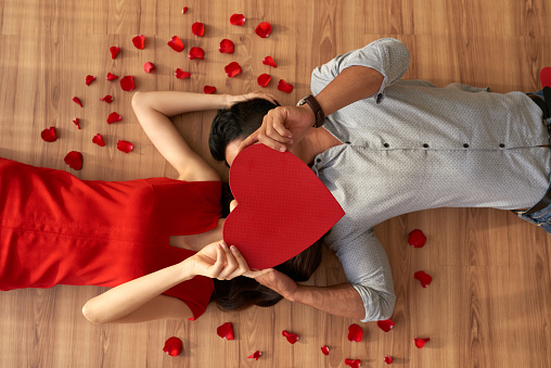 Directly above view of loving young couple covering their faces with heart shaped piece of paper and kissing while lying on floor, red rose petals scattered everywhere