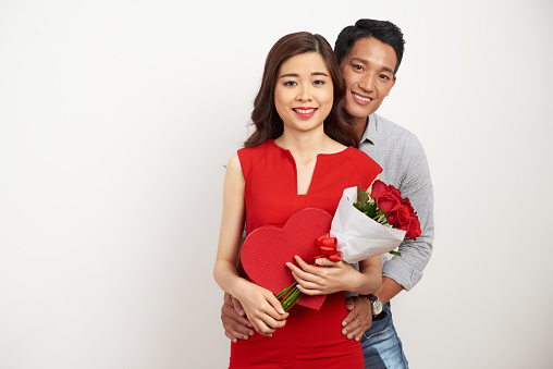 Group portrait of cheerful Asian couple looking at camera with wide smiles while celebrating Valentines Day together, pretty woman holding bouquet of red roses and heart shaped gift box in hands