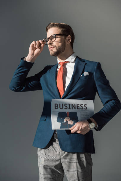 istock handsome young man holding newspaper and adjusting eyeglasses isolated on grey 905956500