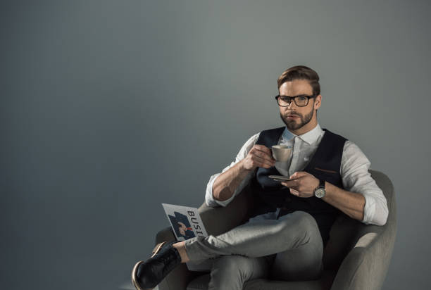 istock handsome young businessman in eyeglasses drinking coffee and looking at camera while sitting with newspaper in armchair isolated on grey 905960730