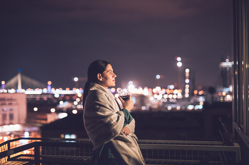Woman standing on the balcony at night wrapped in a blanket and looking on a city view