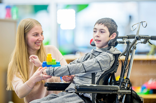 A boy in a wheelchair is indoors in his classroom. He is being assisted by his classroom helper. They are playing with colorful plastic blocks together.