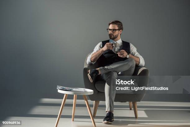 Bel Giovane Elegante Seduto In Poltrona E Distorsi Di Guardare Mentre Beve Caffè - Fotografie stock e altre immagini di Uomini