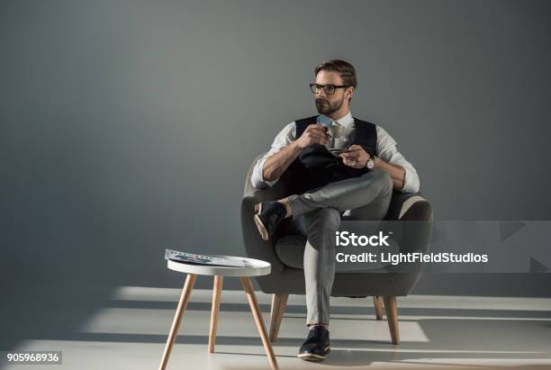 Handsome Stylish Young Man Sitting In Armchair And Looking Away While Drinking Coffee Stock Photo - Download Image Now