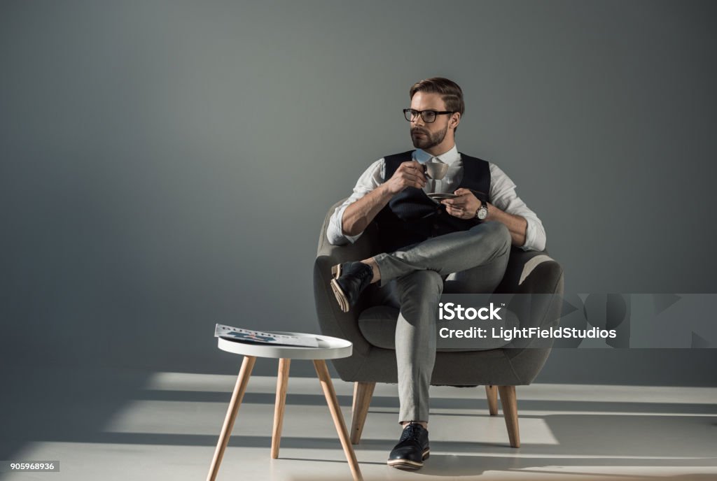 Lindo elegante jovem sentado numa poltrona e desviar o olhar enquanto bebia café - Foto de stock de Homens royalty-free