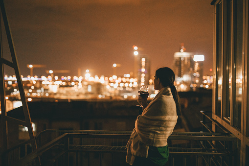 Woman standing on the balcony at night wrapped in a blanket and looking on a city view