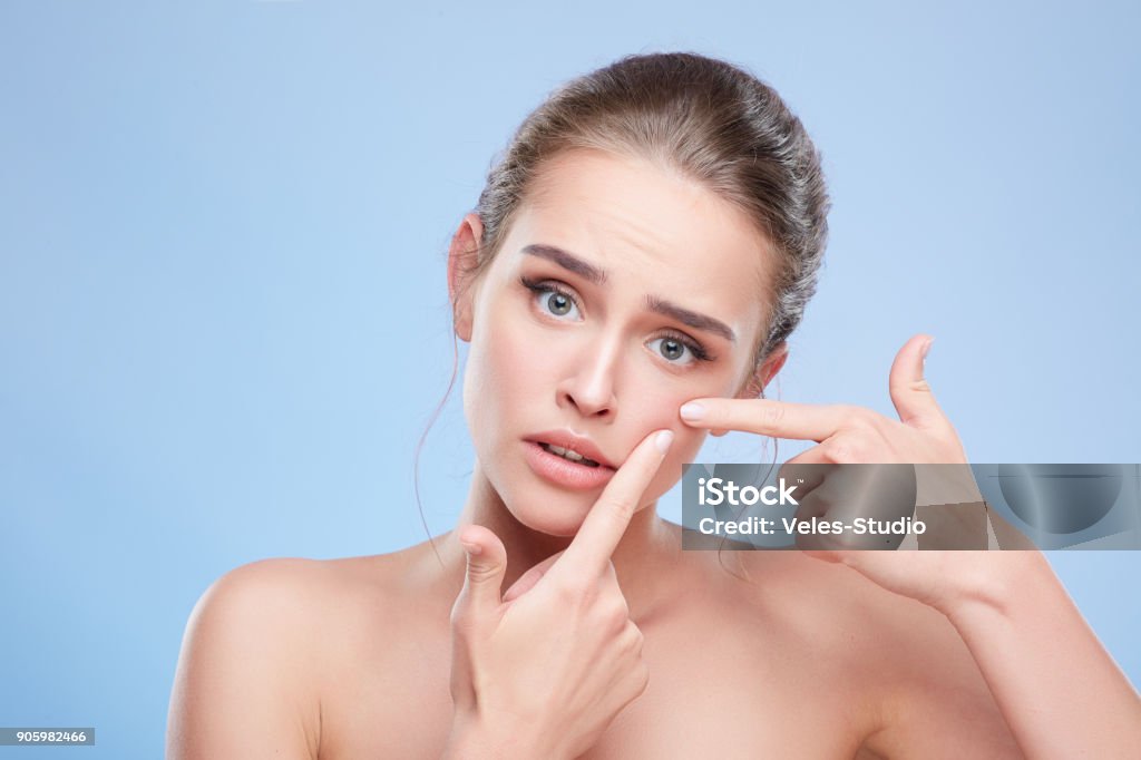 Disappointed girl looking at camera Beauty concept, head and shoulders of young woman with natural makeup. Disappointed girl looking at camera sadly and pressing out pimple on skin. Studio, indoors Acne Stock Photo