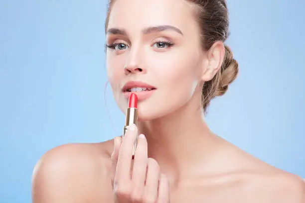 Beauty concept, head and shoulders of young woman with natural makeup. Closeup of girl looking aside and painting lips with scarlet lipstick. Studio, indoors