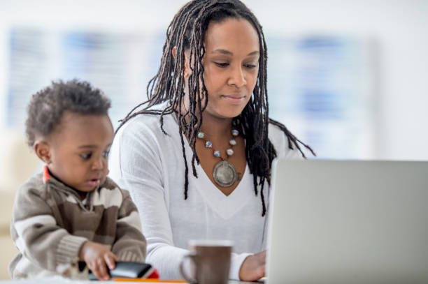 Working Mother A woman of African descent is at home with her baby boy. She is working from home on her laptop. Her son is playing with a phone beside her. Distracted stock pictures, royalty-free photos & images