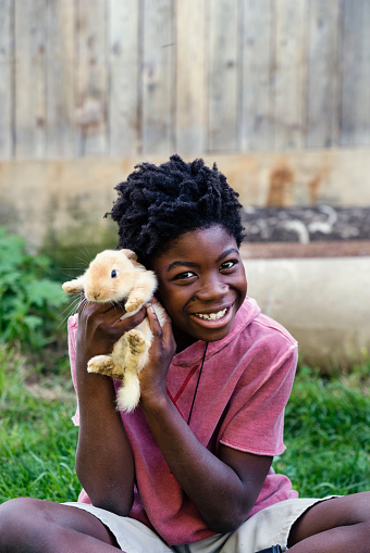 Cute african-american boy holding a beige baby rabbit on her lap, sitting outside near an old barn in springtime. He is wearing a pink hoodie and is looking at camera with a big smile. Vertical waist up outdoors shot with copy space. This was taken in Quebec, Canada.