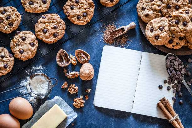 Top view of a kitchen table table filled with chocolate chips cookies and ingredients for preparing and baking cookies wit an open blank cookbook. Some baked cookies are on a cooling rack. Ingredients includes are flour, eggs, chocolate chips, brown sugar and butter. Some kitchen utensils complete the composition. Predominant color is brown. Low key DSRL studio photo taken with Canon EOS 5D Mk II and Canon EF 100mm f/2.8L Macro IS USM