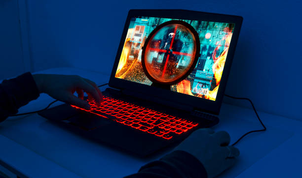 Gaming laptop stands on a table. It is night and the keyboard lights up with red colors, and a computer mouse is connected to the pc. The screen shows a video game with a soldier firing a rifle. The image on the screen is the photographer's work.