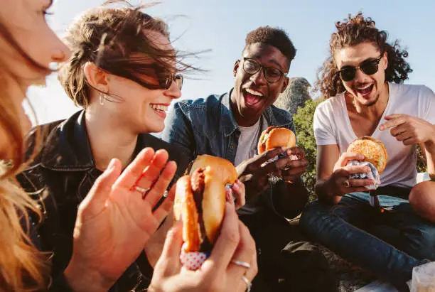 Group of friends sitting on mountain top eating burger. Excited young men and women enjoying and partying outdoors.