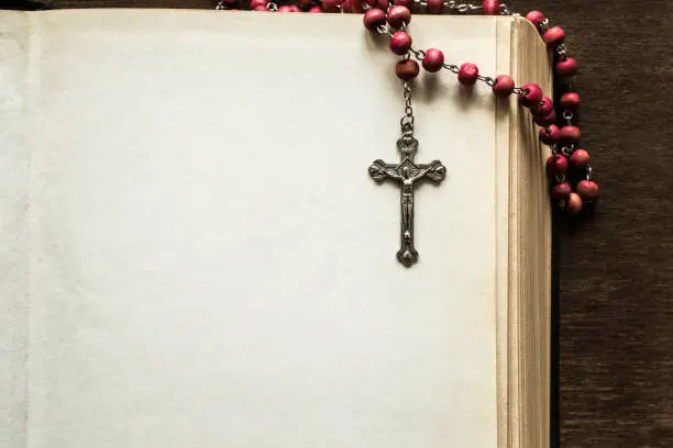 Opened old thick bible with rosary beads on the brown table in the quiet, dark atmosphere. Prayer time on sunday. Empty place for text. Religion concept.
