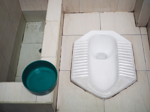 Old Thai local traditional, countryside style white ceramic squatty potty toilet room, dirty white tile floor, wall, water basin and green bucket for cleaning, perspective top view