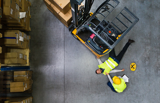 An accident in a warehouse. Woman with smartphone and her colleague lying on the floor next to a forklift. Aerial view.An accident in a warehouse. Woman running towards her colleague lying on the floor next to a forklift.