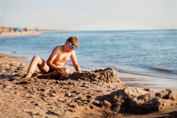 A boy in swimming goggles is having fun while playing with sand in the evening on a beach.