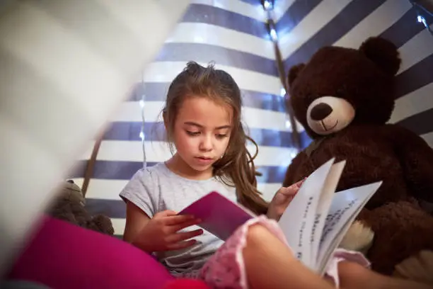 Cropped shot of a little girl reading a book with her teddybear in a tent at home