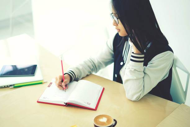 istock Skilled female trainee concentrated on writing publication noting ideas with pen sitting in coworking space near copy space for advertising, clever teen pupil doing homework pondering on task 906655314