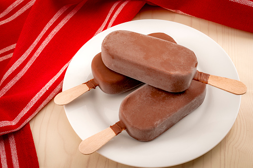 Bars of vanilla ice cream on a stick, with a chocolate coating in a rustic setting and a red towel in the background