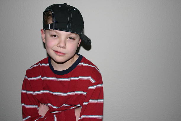 Little, cheeky blond boy with curly hair, lying on the floor in front of a white studio background. Wearing a cap and looking at the camera, giving thumbs up to the camera. Full length image.
