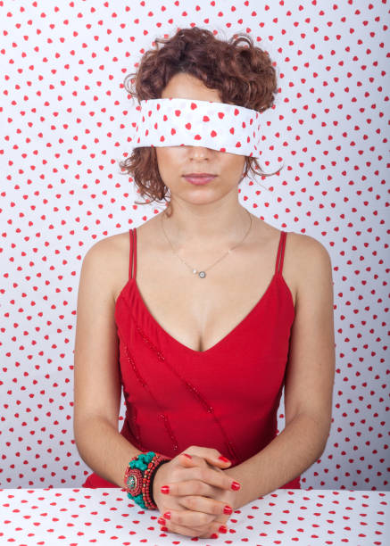 Blindfolded Beautiful Adult Woman With Red Hair Portrait of mesmerized blindfolded adult woman wearing a red dress. Her hair is long and red. The background is red and white heart shape dotted fabric. Shot in studio with a full frame DSLr camera. red blindfold stock pictures, royalty-free photos & images
