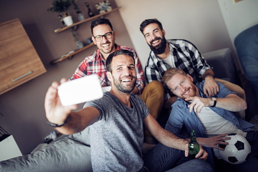 Cheerful group of friends having fun at home,taking selfie and watching football game.