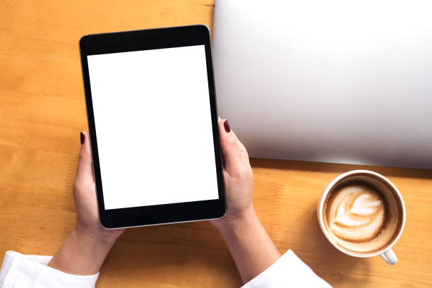 istock Top view mockup image of woman's hands holding black tablet pc with white blank screen and laptop , coffee cup on wooden table background 906734218