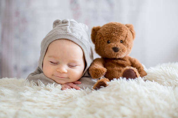 Sweet baby boy in bear overall, sleeping in bed with teddy bear Sweet baby boy in bear overall, sleeping in bed with teddy bear stuffed toys, winter landscape behind him bonnet hat stock pictures, royalty-free photos & images