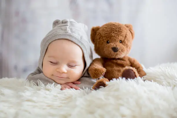 Photo of Sweet baby boy in bear overall, sleeping in bed with teddy bear