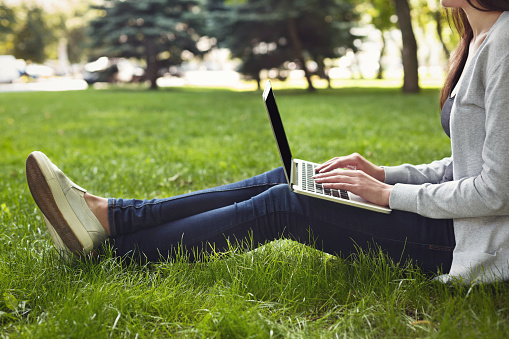 Young unrecognizable woman sitting outdoors on grass with laptop, typing, surfing internet, preparing for exams. Technology, communication, education and remote working concept, copy space