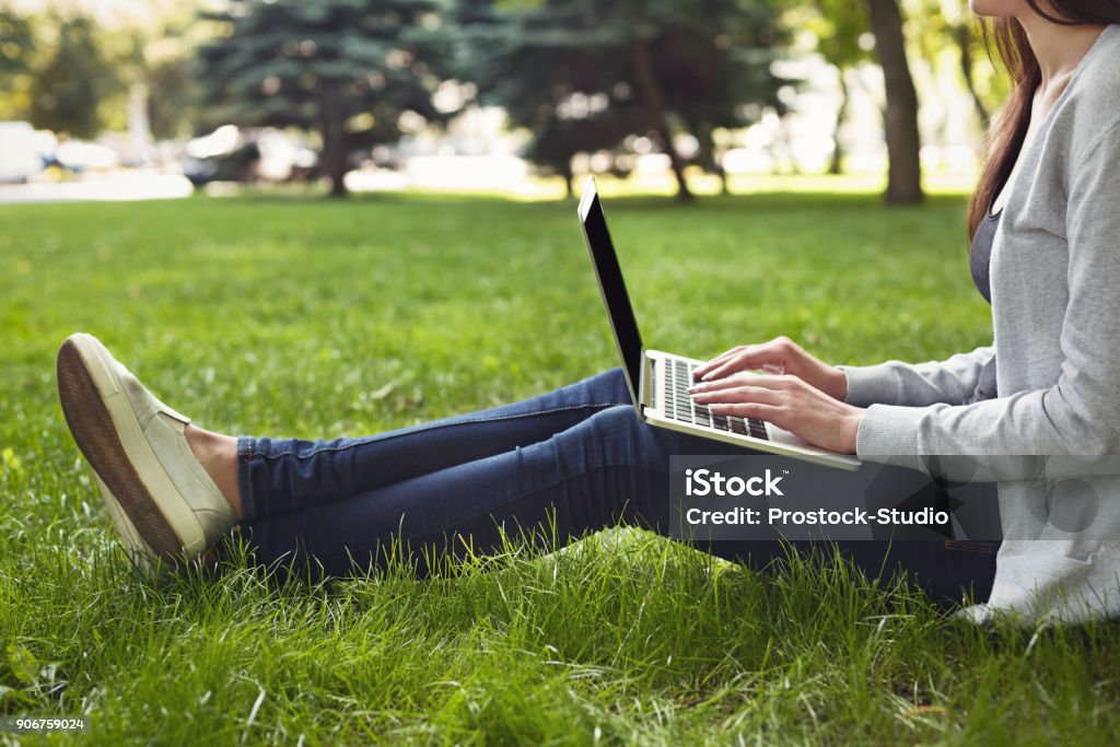 Young pensive woman using laptop in park Young unrecognizable woman sitting outdoors on grass with laptop, typing, surfing internet, preparing for exams. Technology, communication, education and remote working concept, copy space Outdoors Stock Photo