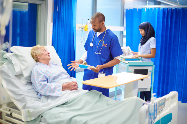 explaining the operation a mid adult doctor chats to a senior patient on the hospital ward , in the background a young female nurse wearing a hijab is tending to the records. pre operative stock pictures, royalty-free photos & images