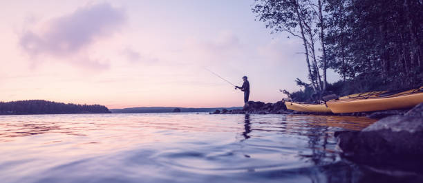 Peaceful fishing at a lake Late evening panorama of a lake with a fisher and two kayaks on the shore. rowing boat fishing stock pictures, royalty-free photos & images