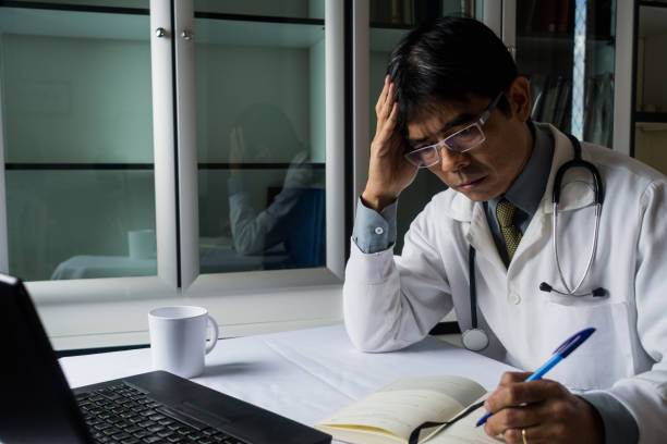 istock Distressed asian doctor sitting at desk working on laptop taking notes having a cup of coffee tea 906908948