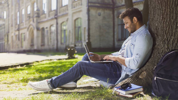 istock Young mixed-race guy using laptop under tree, smiling at good news, emotions 906952376