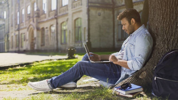 istock University student under tree, using laptop looking surprised, upsetting news 906952622
