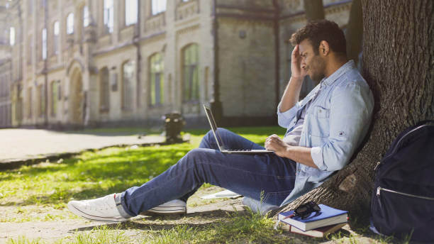 istock College student sitting under tree with laptop looking upset disappointing news 906952880