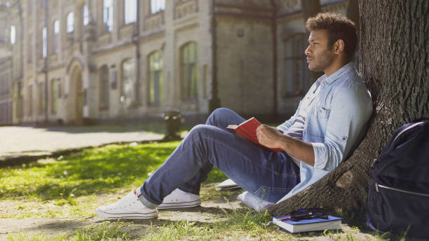 istock Male university student sitting under tree, reading book, memorizing information 906953694