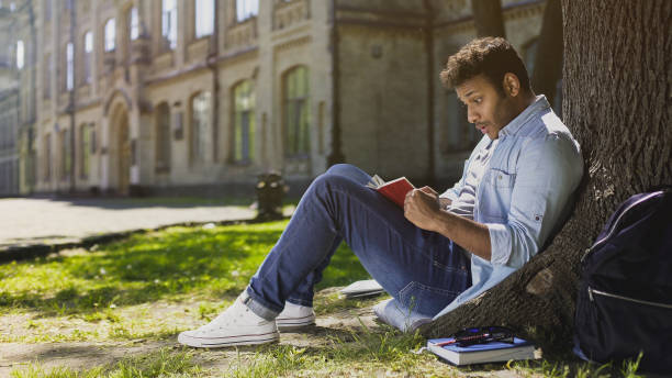 istock Multinational young male sitting under tree, reading gripping book, surprised 906953880