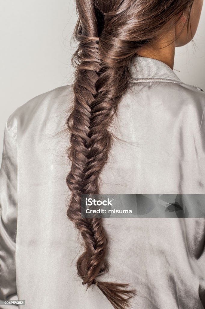 Beauty and fashion. Portrait of young woman with a long braided pigtail, dressed in a metallic colored bomber jacket, back view. Braided Hair Stock Photo