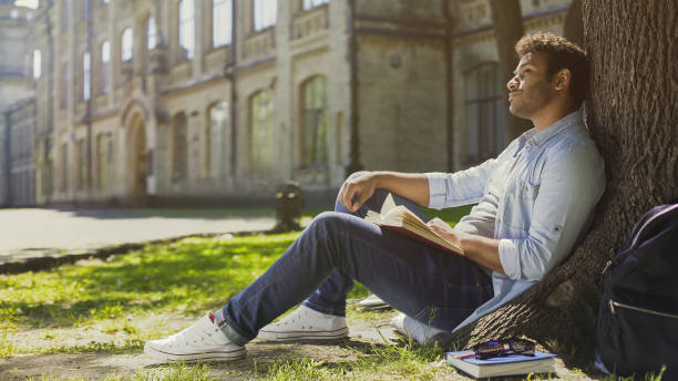 istock Young male sitting under tree with book looking around, having pleasant thoughts 906954578