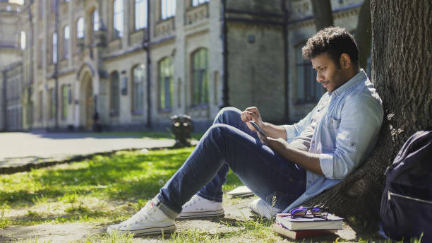 istock Multinational male sitting under tree using phone, reading newsfeed on gadget 906954990