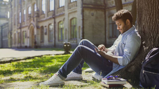 istock Young man sitting under tree in park, using mobile phone and smiling, good news 906957204