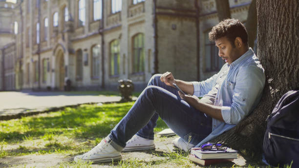 istock Surprised guy sitting under tree, using mobile phone, reacting to newsfeed 906959080