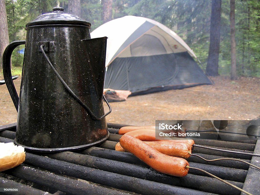 Cooking Food On A Camping Trip A kettle and hot dogs on a smoking grill, with a camping tent and trees in the background. Focus on the kettle and hot dogs. Barbecue - Meal Stock Photo