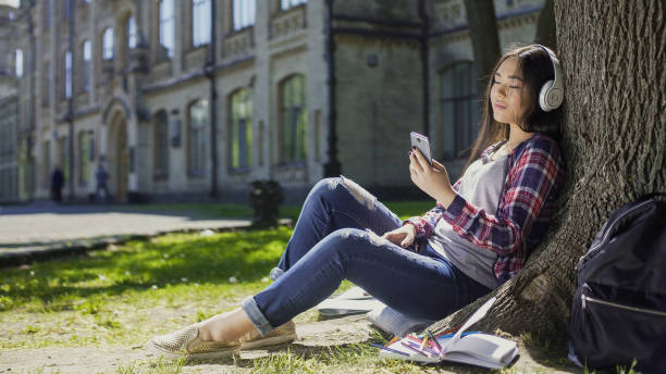 istock Mixed-race girl under tree in headphones, scrolling phone screen, choosing song 906964094