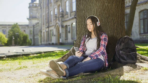 istock Multinational girl sitting under tree in headphones, using phone, choosing song 906964528