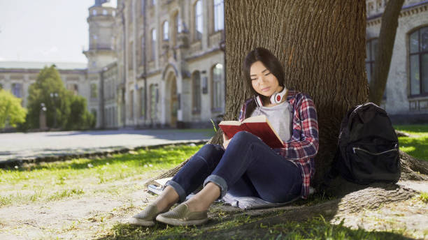 istock Mixed female sitting under tree, reading favorite book, gripping plot, engrossed 906965230
